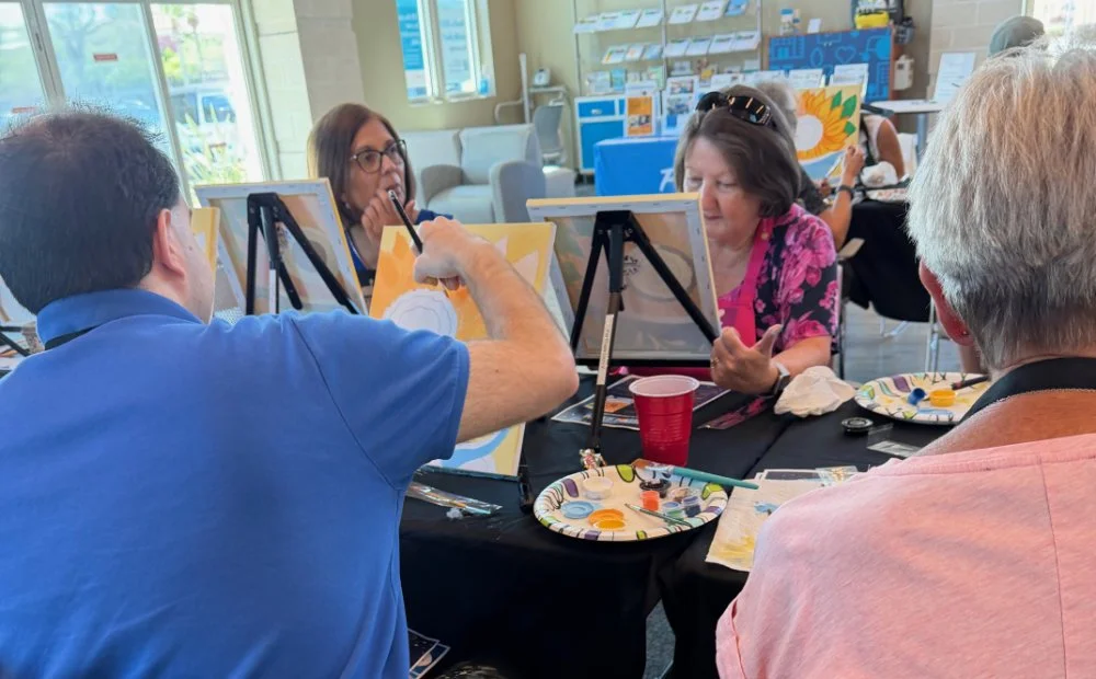 People participating in a group painting activity at a table in a well-lit room, with canvases, paints, and brushes.