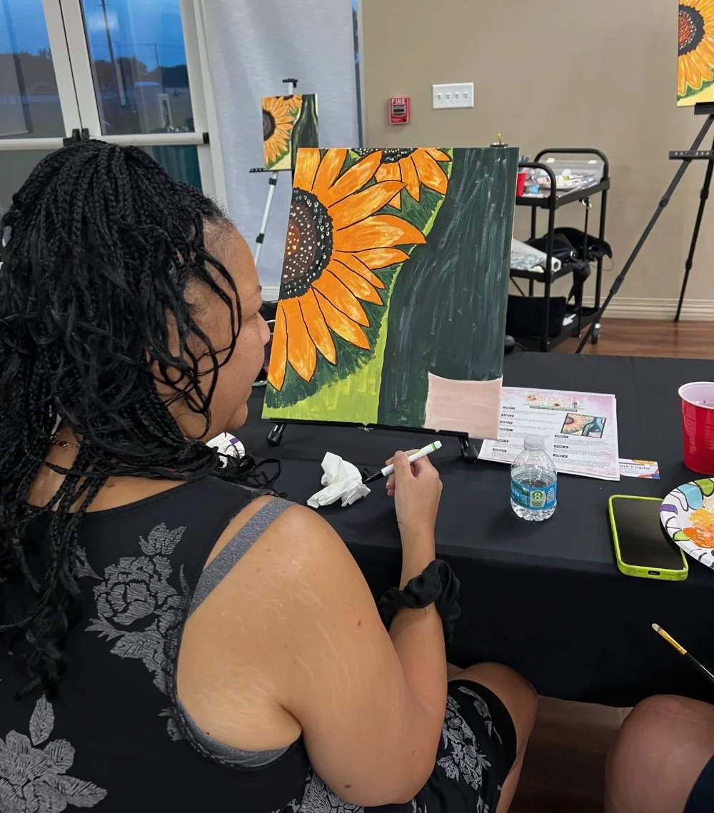 A woman with braided hair painting a sunflower on a canvas in a painting class. There are art supplies, a water bottle, and a snack on the table, with other canvases and easels in the background.