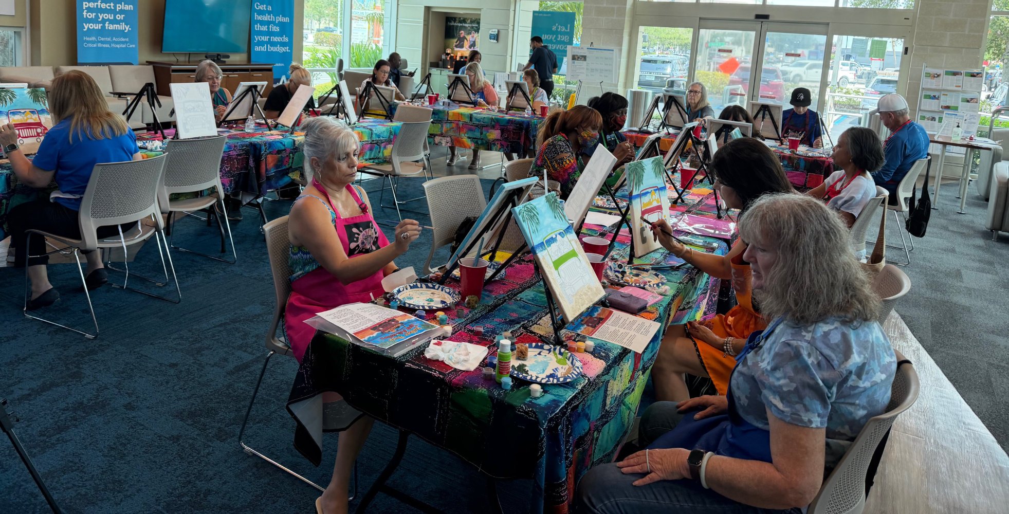 A group of people participating in a painting class at a studio, sitting at tables with colorful tablecloths and painting supplies, creating tropical beach scenes on canvases.