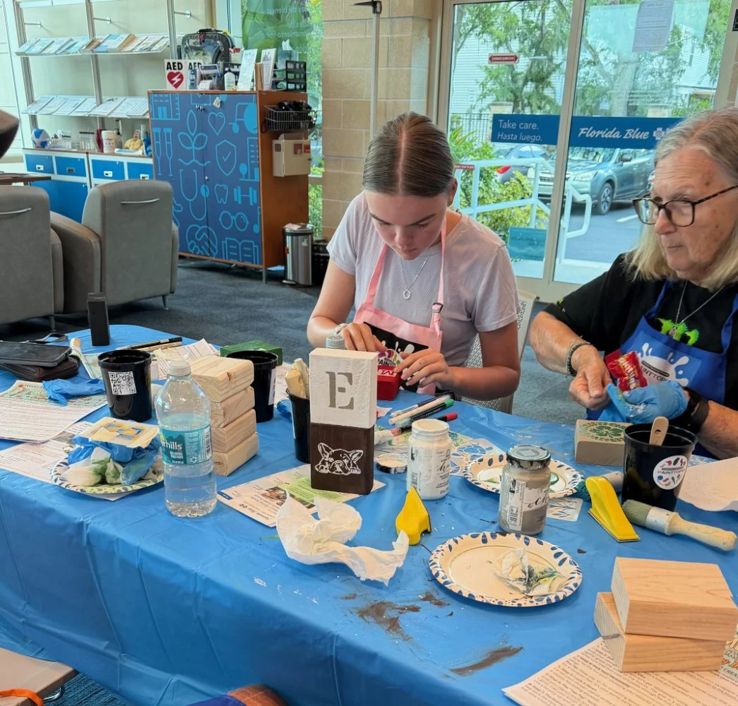 Two women sit at a craft table covered with a blue plastic tablecloth, working on wooden art projects. The table has paint jars, brushes, paper plates with paint, and various craft supplies. One woman wears glasses and a black apron, while the younge