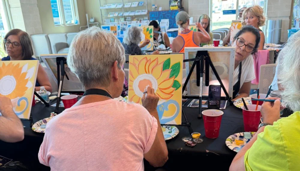 Group of people painting sunflower pictures on easels at a social art event in a bright room.