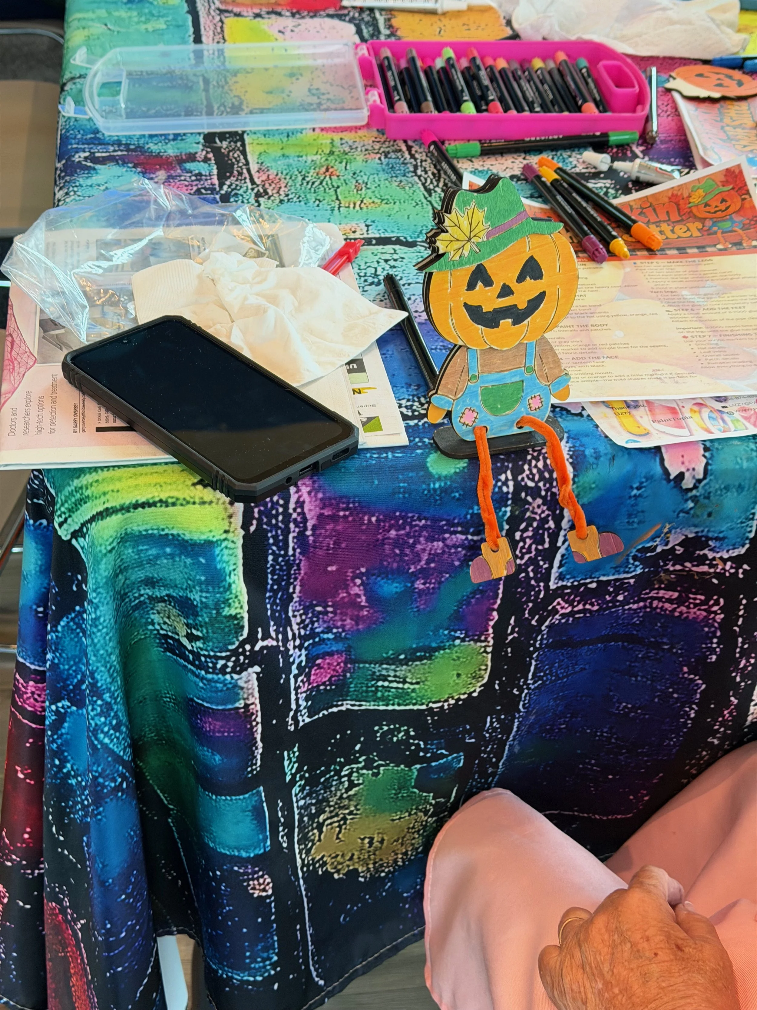 A colorful tablecloth with a Halloween-themed craft project including a paper jack-o'-lantern figure with a green hat decorated with a yellow leaf and painted face, surrounded by various markers, paper, tissues, a smartphone, and other craft supplies