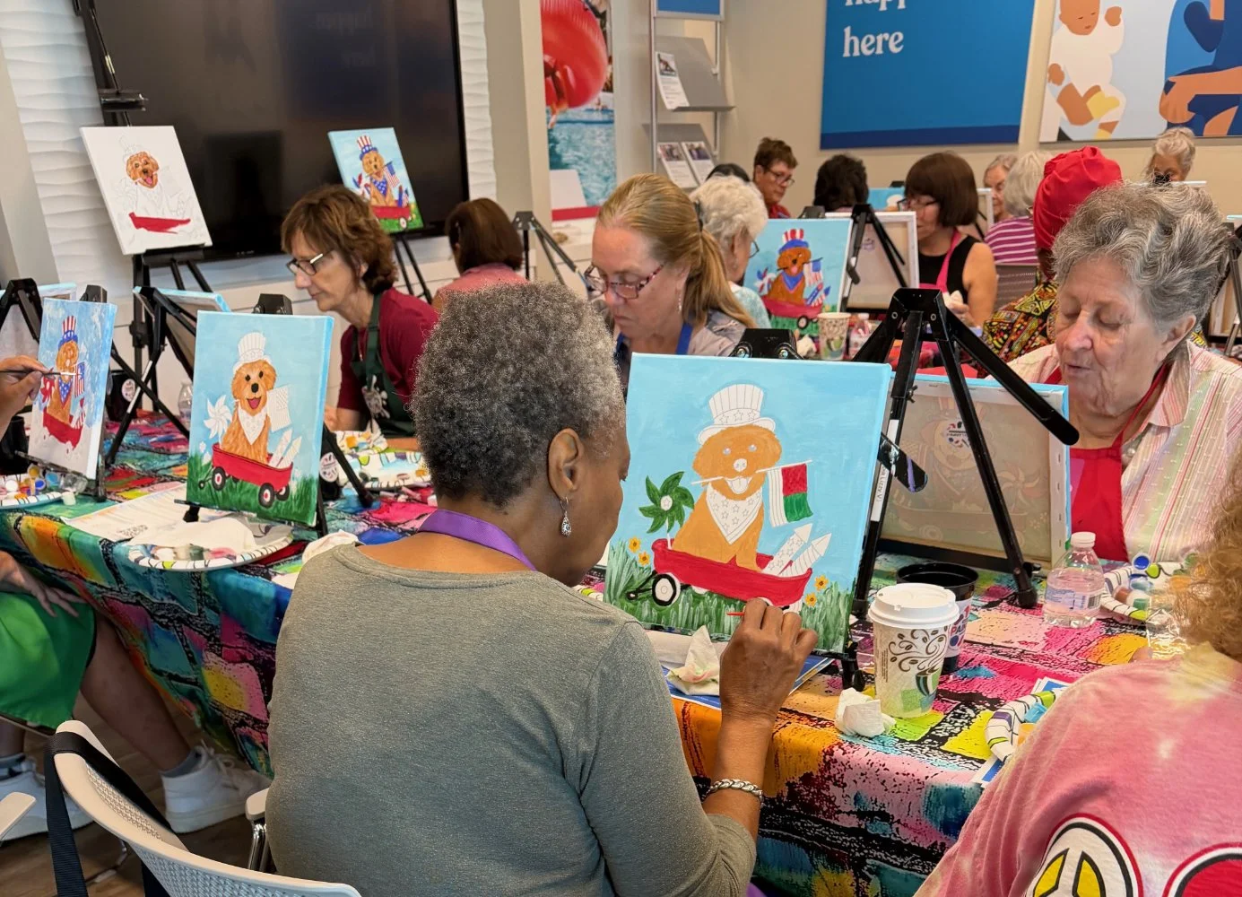 Older women participating in a paint and sip class, painting pictures of a golden retriever with a chef's hat on small canvases, sitting at tables with colorful tablecloths, in a group setting.