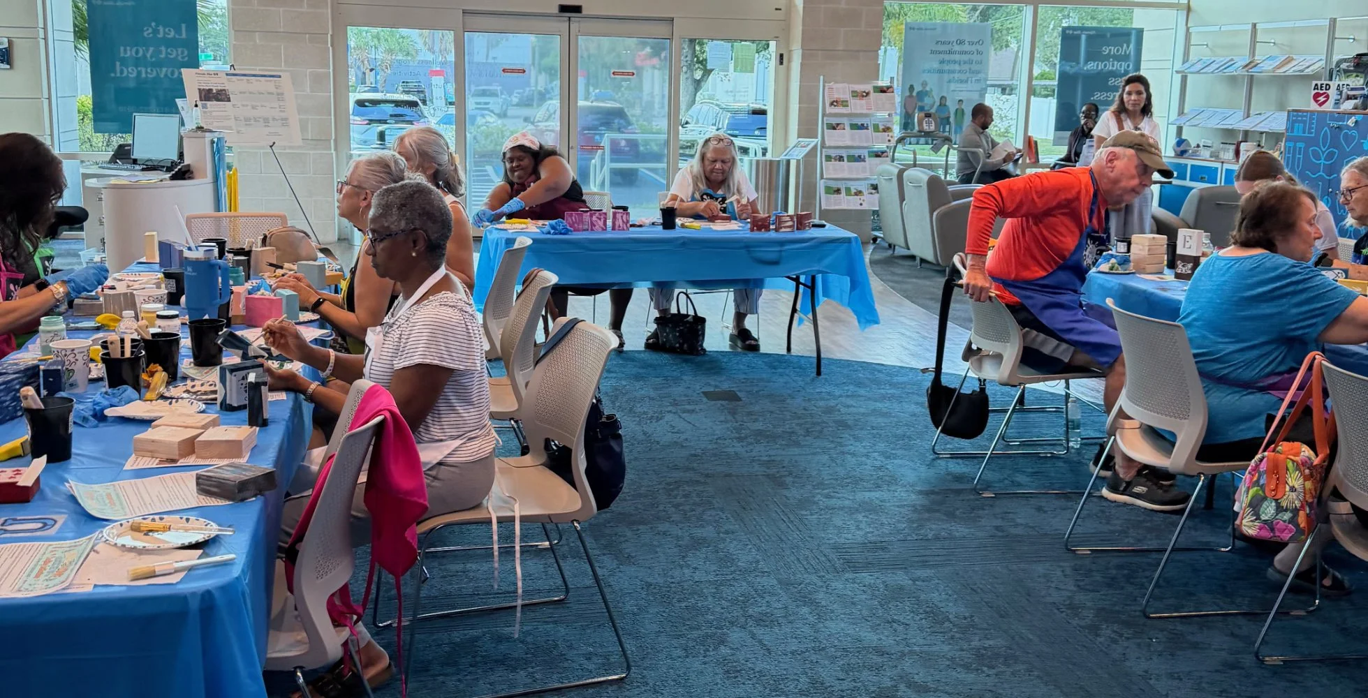 People participating in a craft event inside a community center or medical facility, with tables covered in blue tablecloths, arts and crafts supplies, and informational brochures in the background.