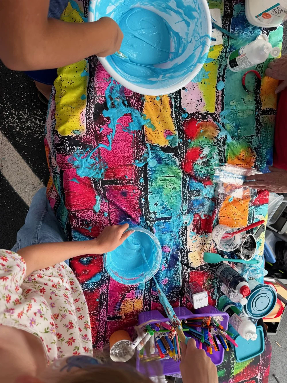 Children engaging in a paint activity on a colorful tablecloth, mixing blue paint in bowls with various art supplies around.