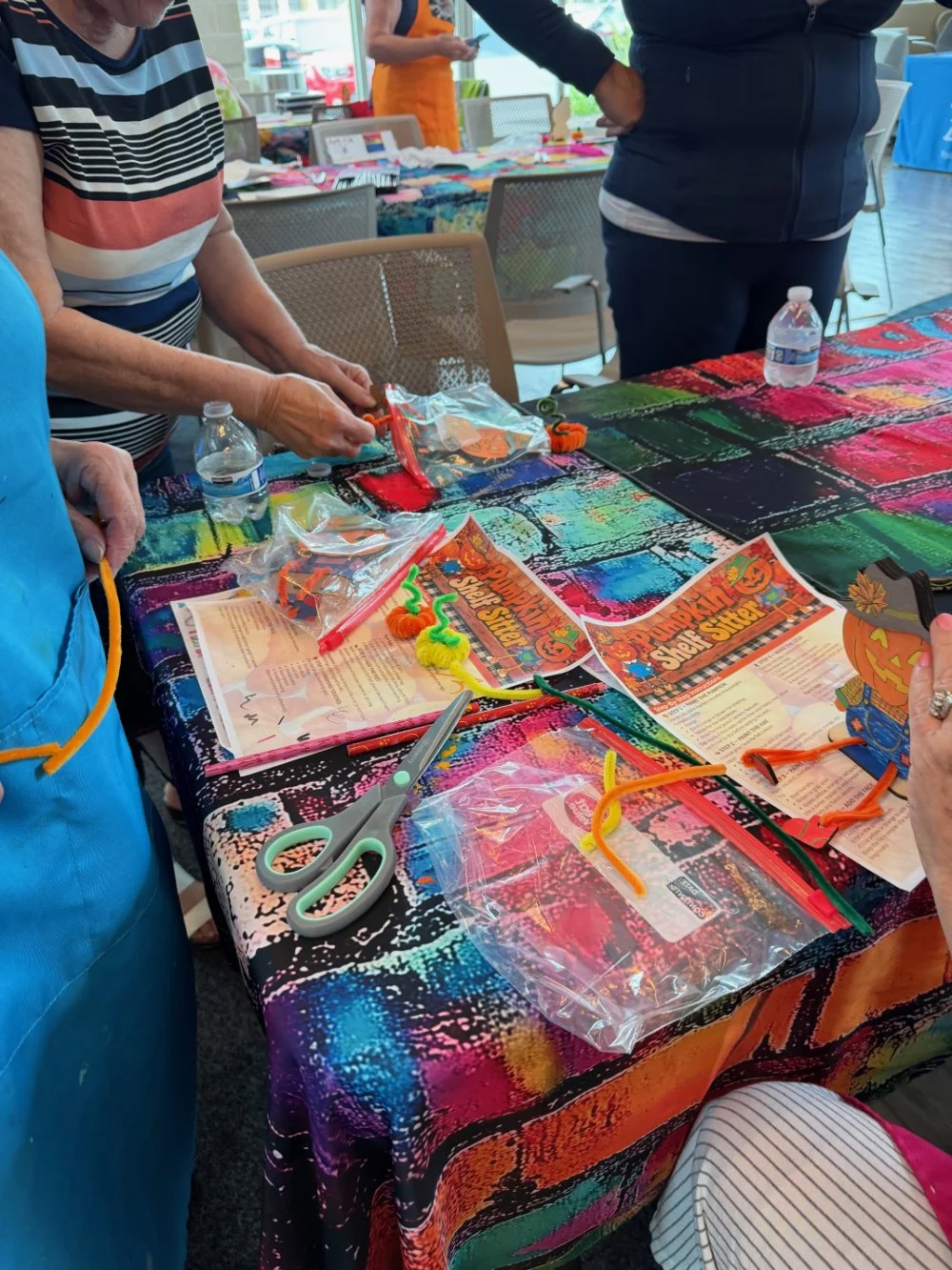 People gathered around a colorful table with Halloween-themed flyers, pipe cleaners, scissors, and plastic bags, preparing craft materials for a Halloween event.