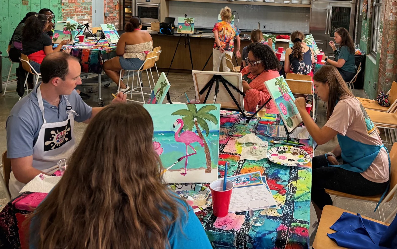People participating in a group painting class, creating tropical-themed artwork with palm trees and flamingos on canvases, at a colorful, artistic studio with art supplies on the tables.