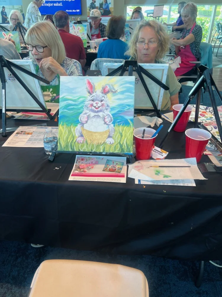 A group of people participating in a painting workshop, with a colorful canvas of a smiling anthropomorphic bunny dressed as a rabbit in the foreground, painting supplies, red cups, and informational pamphlets on the table.