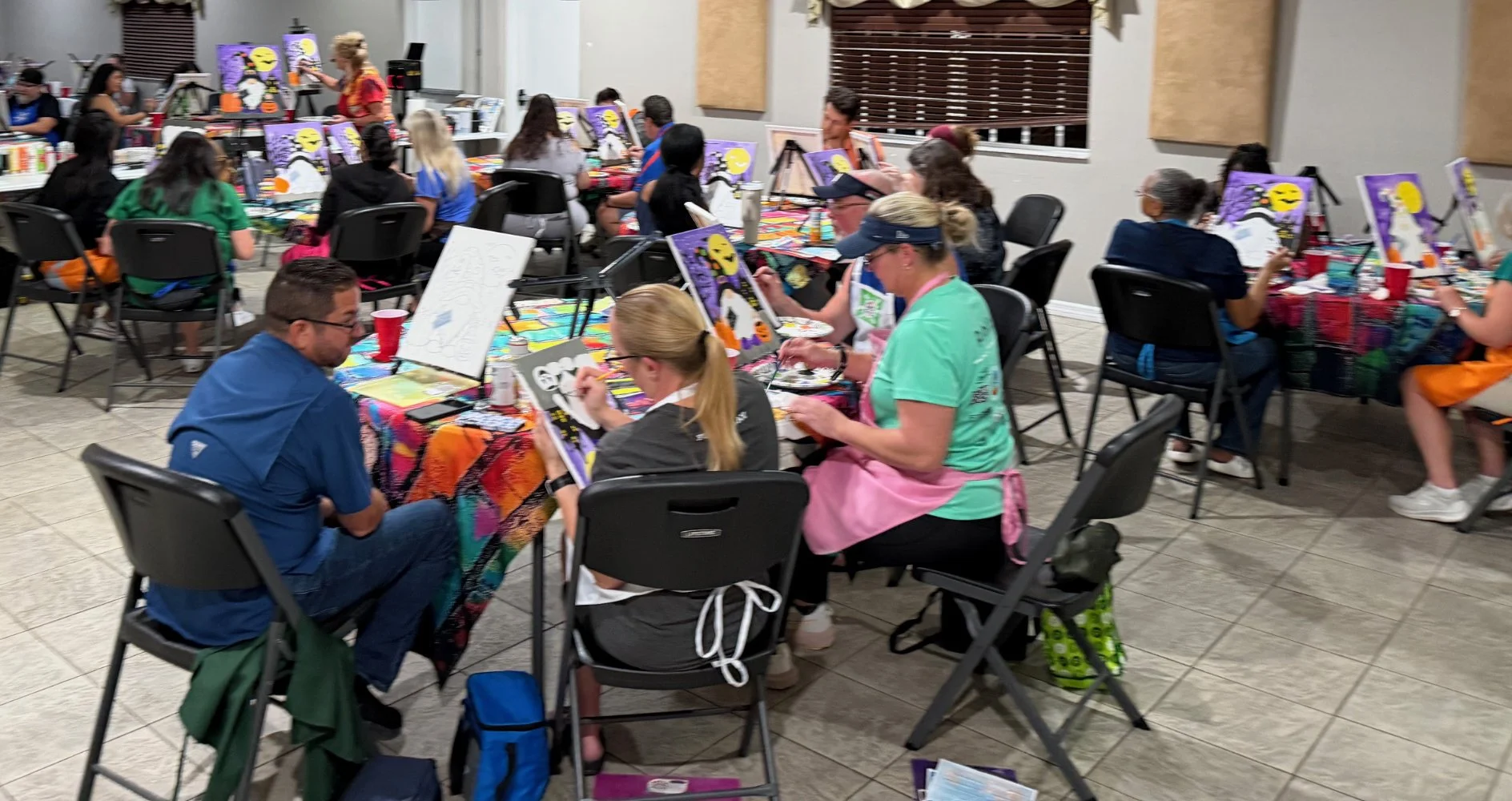 A group of people at a painting class in a room, painting Halloween-themed pictures with a woman instructor guiding them. The room has tables with colorful tablecloths, art supplies, and canvases. Participants are wearing aprons and some are wearing 