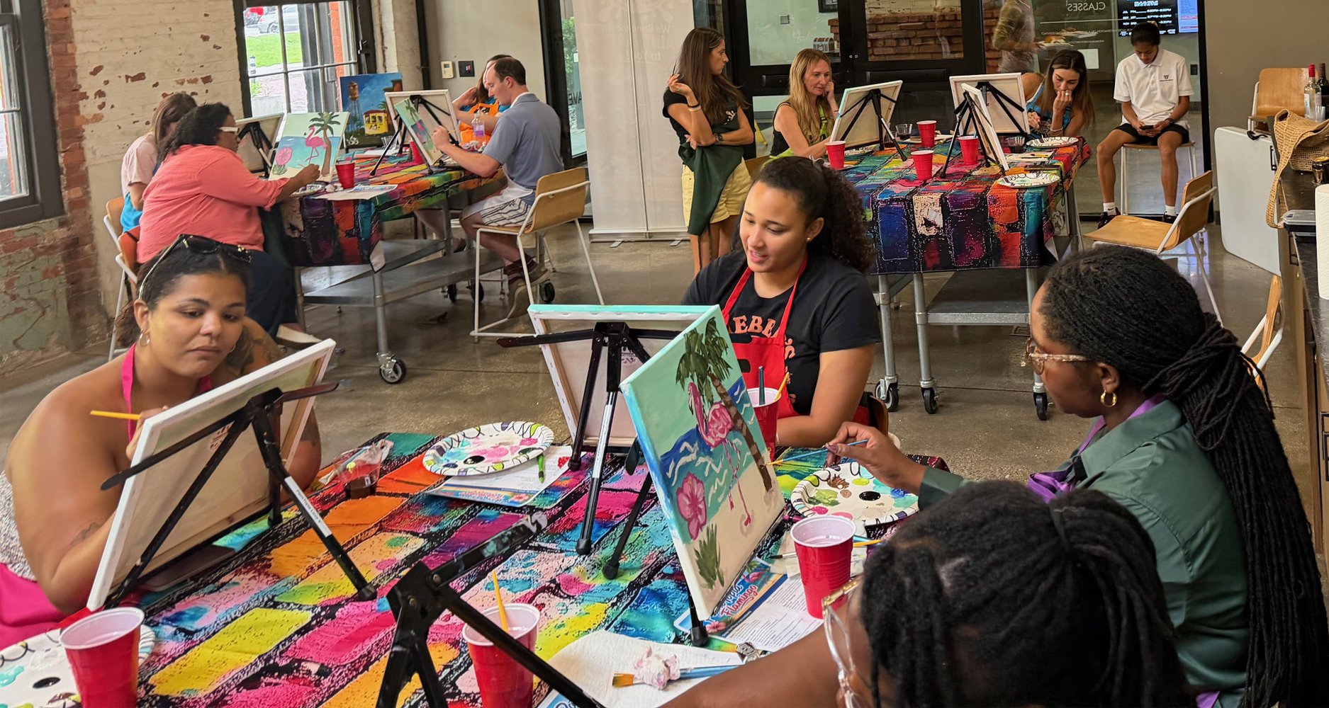 Women and girls participating in a group painting event in an art studio with colorful tablecloths, easels, and paintings.