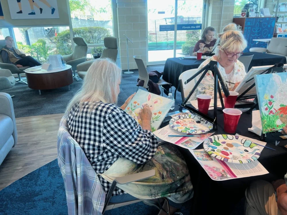 Older women participating in a painting class at a community space, with paintings, brushes, and red cups on the table.