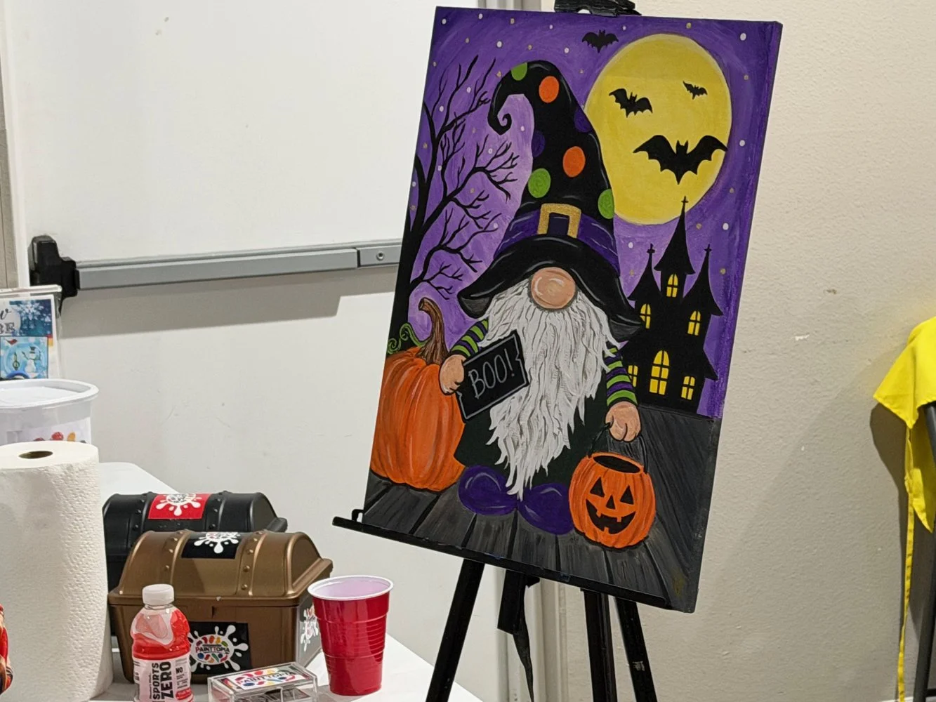 A colorful Halloween-themed painting of a gnome with a tall, polka-dotted hat, holding a pumpkin-shaped bucket and a sign that says 