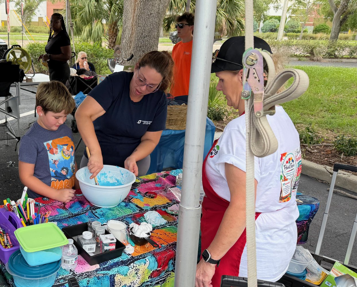 People at an outdoor craft booth, with a woman and young boy mixing blue paint in a large bowl, surrounded by colorful art supplies, with others and trees in the background.