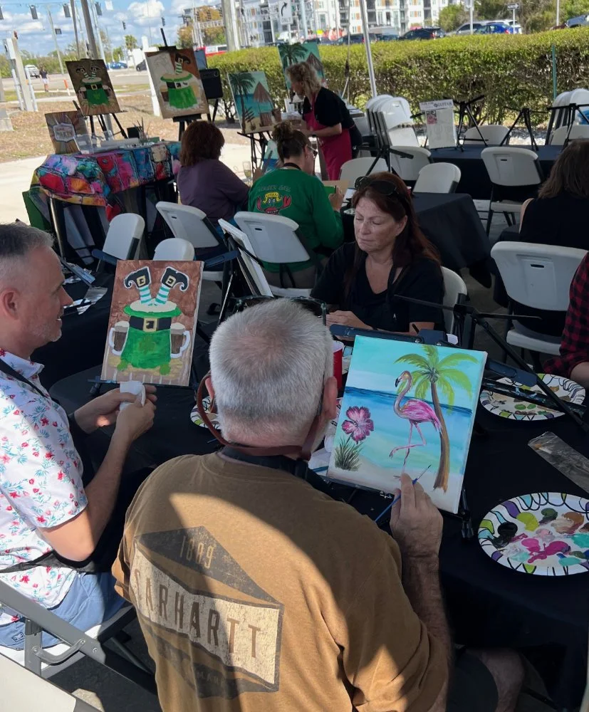 People participating in a painting event outdoors, with various paintings of tropical themes and humorous characters, sitting at tables with art supplies.