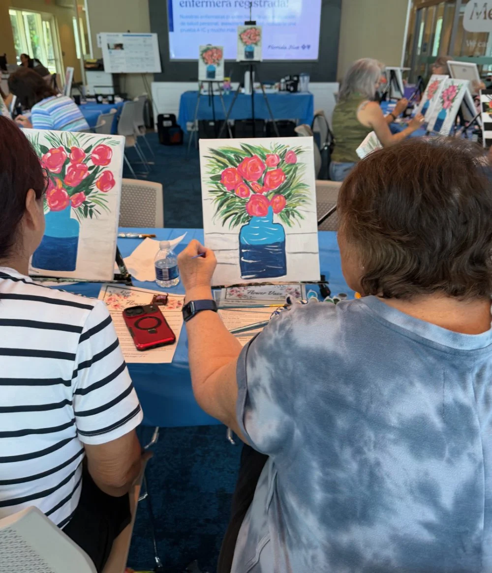 Women in a painting class creating artwork of flowers in a blue vase, with completed paintings of similar floral arrangements on easels. The room has multiple participants, tables with blue tablecloths, and viewers watching a presentation at the fron