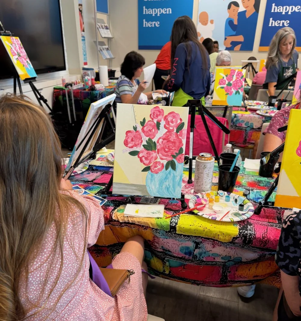 A woman painting pink roses in a vase at a black easel during a group painting session at a colorful table.