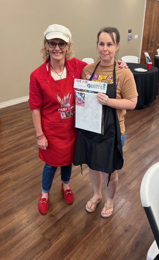 Two women standing indoors, one with short blonde hair and glasses wearing a red apron and hat, the other with brown hair in a ponytail wearing a brown T-shirt and black apron, holding an art kit.