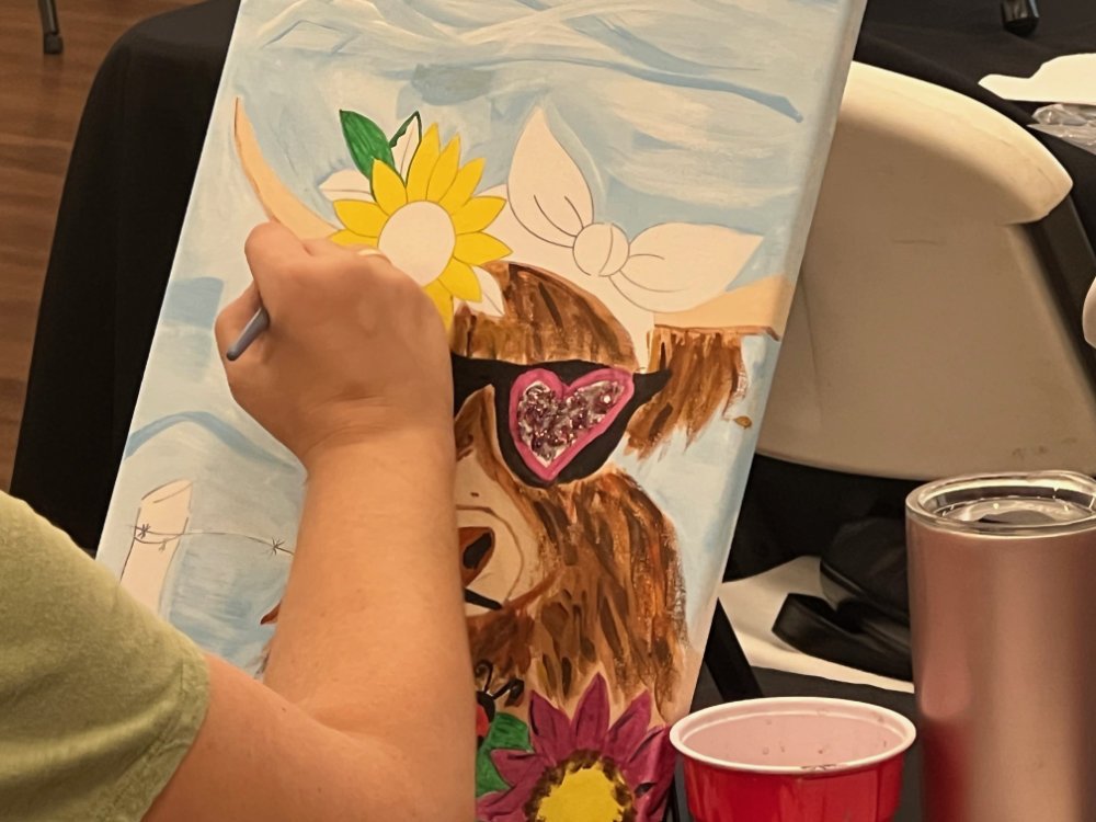 Person painting a colorful artwork featuring a sunflower, a butterfly, a bear's face with a heart-shaped nose, and other floral details.