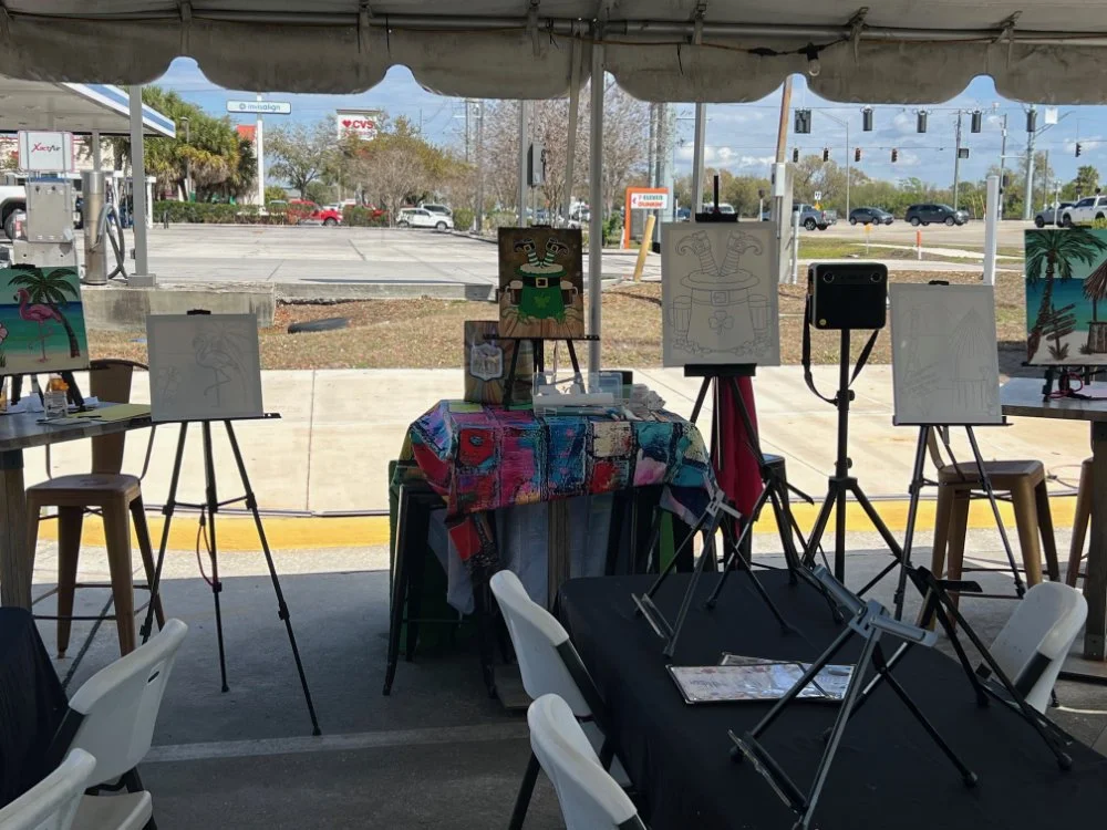 Empty art exhibit booth with paintings on easels and tables under a tent, with a parking lot and street visible in the background.
