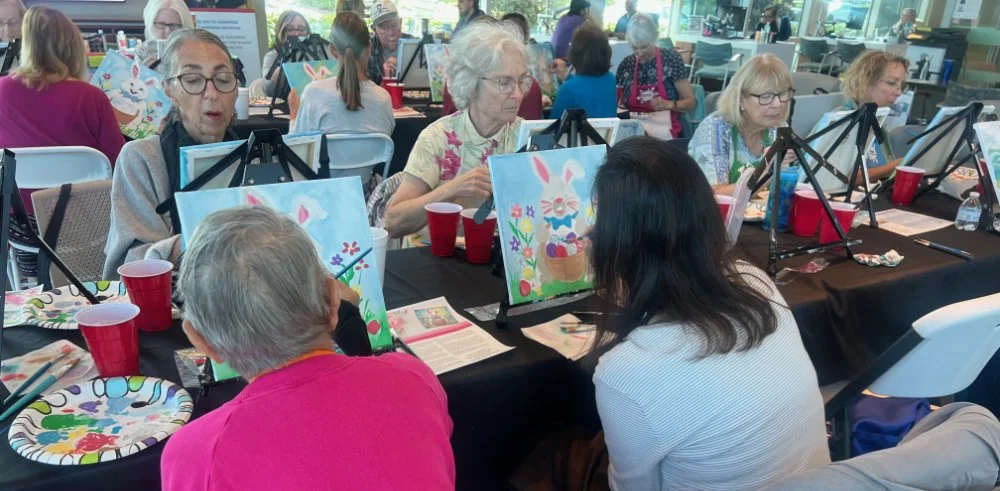Group of elderly women and women in middle age participating in a painting class, sitting at tables with easels, paintings of bunnies and bunnies' eggs, cups, and art supplies.