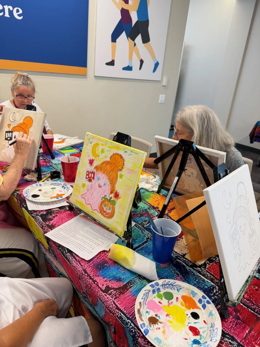 People participating in a painting class, working on individual artwork at a colorful table with painting supplies and protective fabrics.