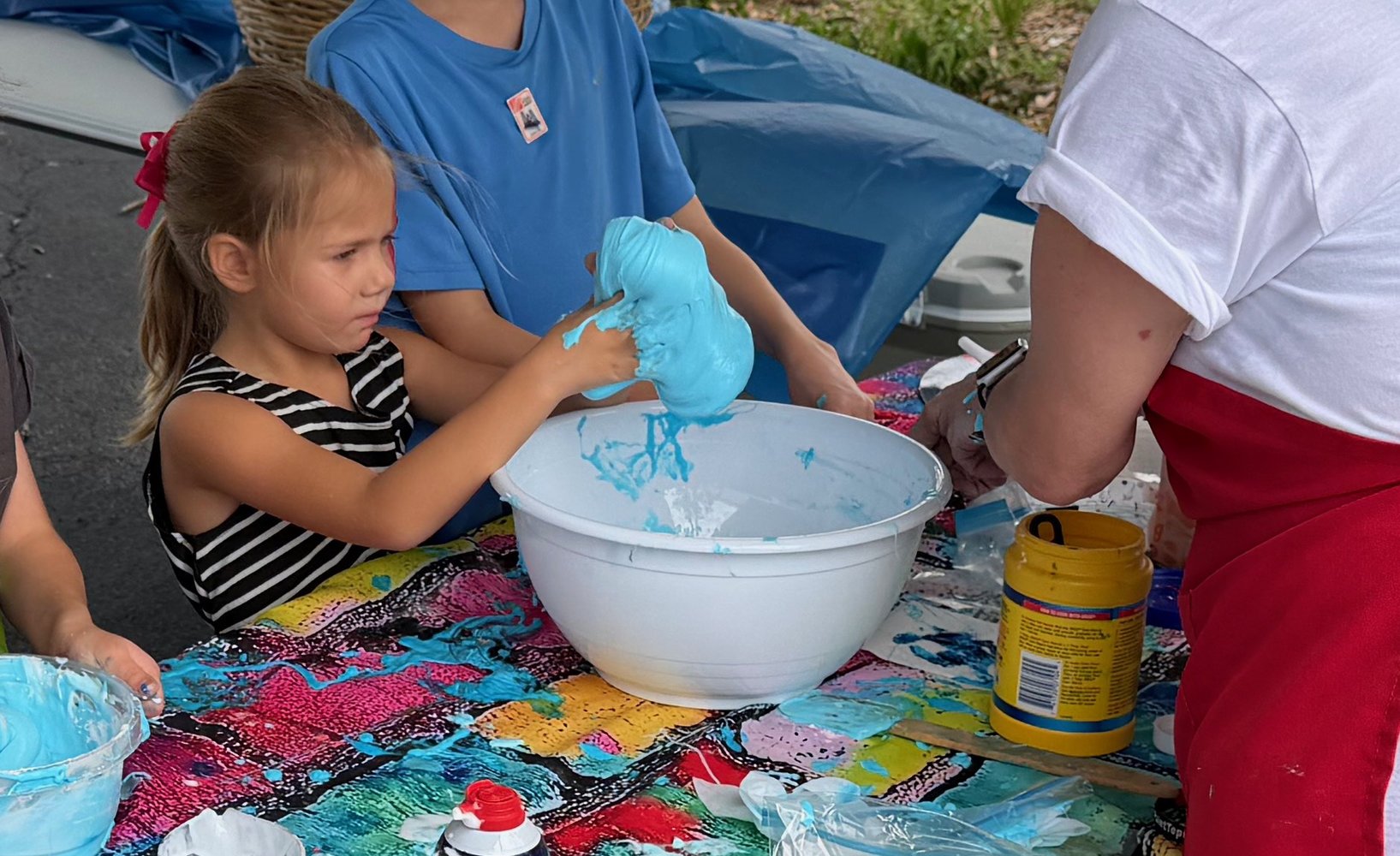 A young girl with a striped shirt is involved in a tie-dye activity, handling a bunch of blue fabric or material with a focused expression. She is sitting at a table covered with a colorful cloth, with various supplies and containers of dye or paint 