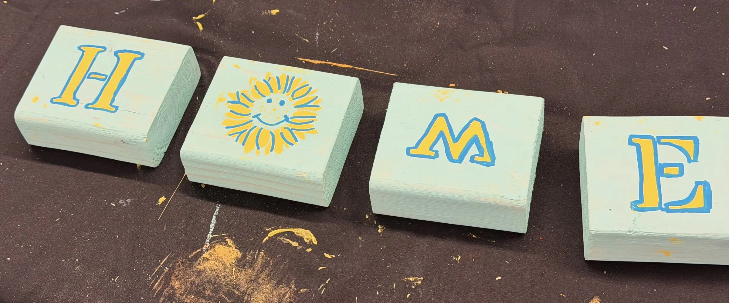 Four painted wooden blocks laid out on a dark surface displaying different designs and letters, including a large yellow H, a smiling sun with blue sun rays, a yellow M with blue outlines, and a yellow 12 with blue outlines.