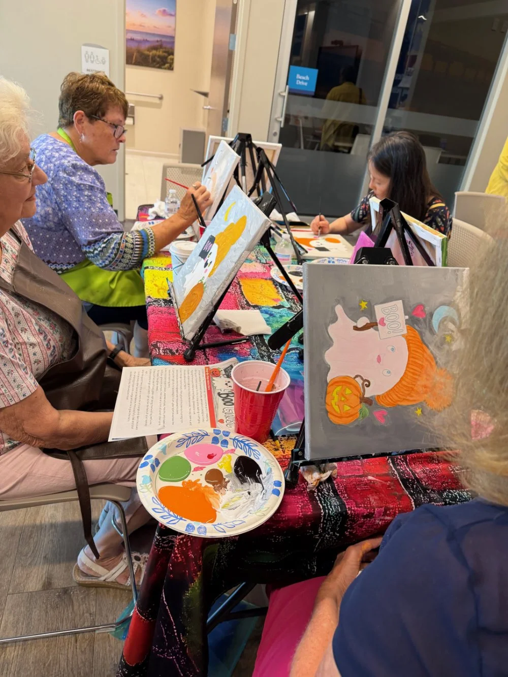 Older women and a young girl painting Halloween-themed pictures on canvases at a table, with painting supplies and colorful decorations.