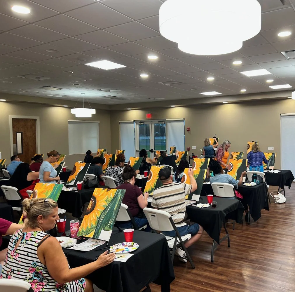 People participating in a group painting class in a well-lit room, each creating a sunflower painting on canvases, with painting supplies and drinks on the tables.