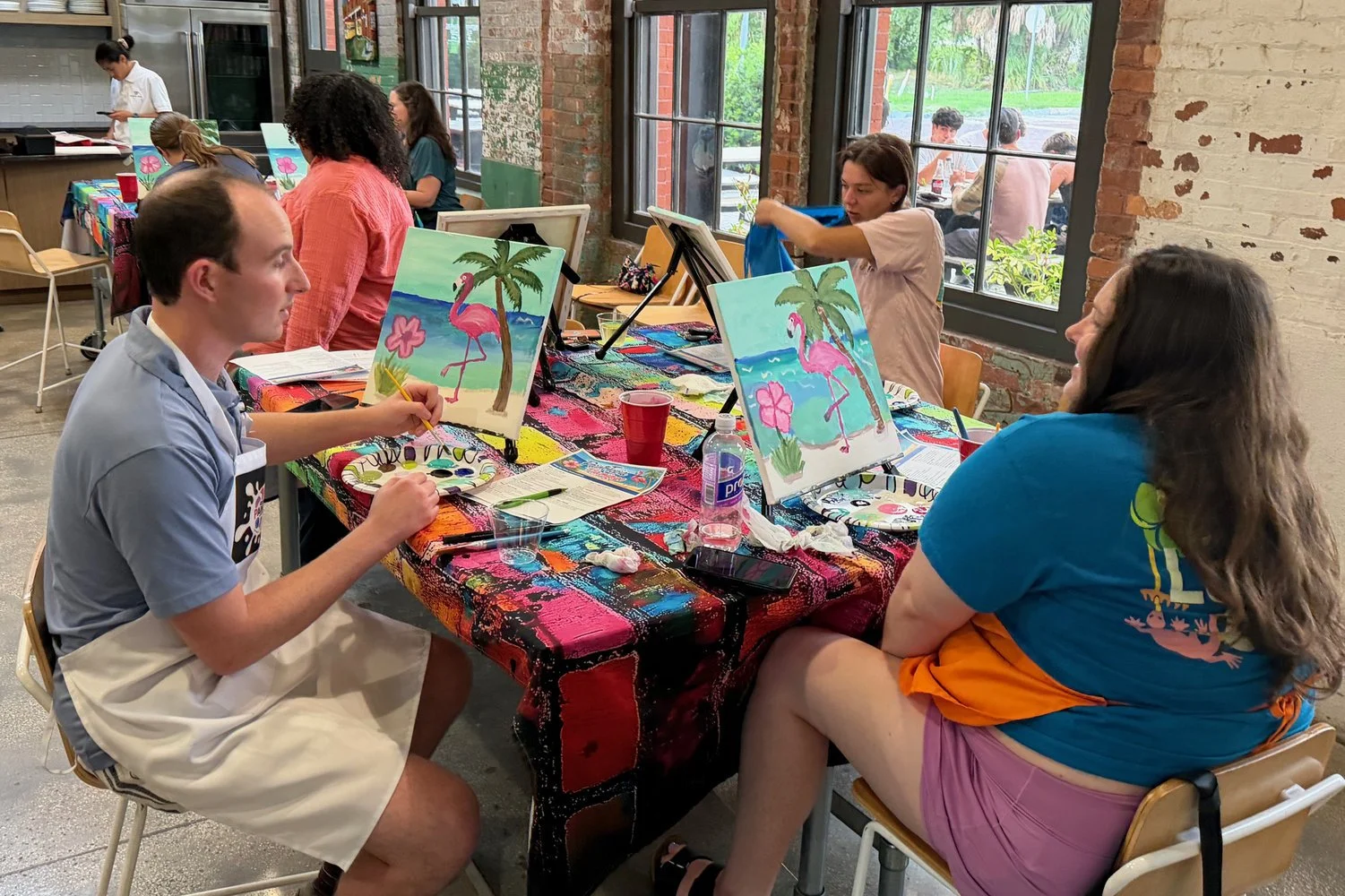People participating in a painting class at a table covered with colorful cloth, working on paintings of pink flamingos on a beach with palm trees.