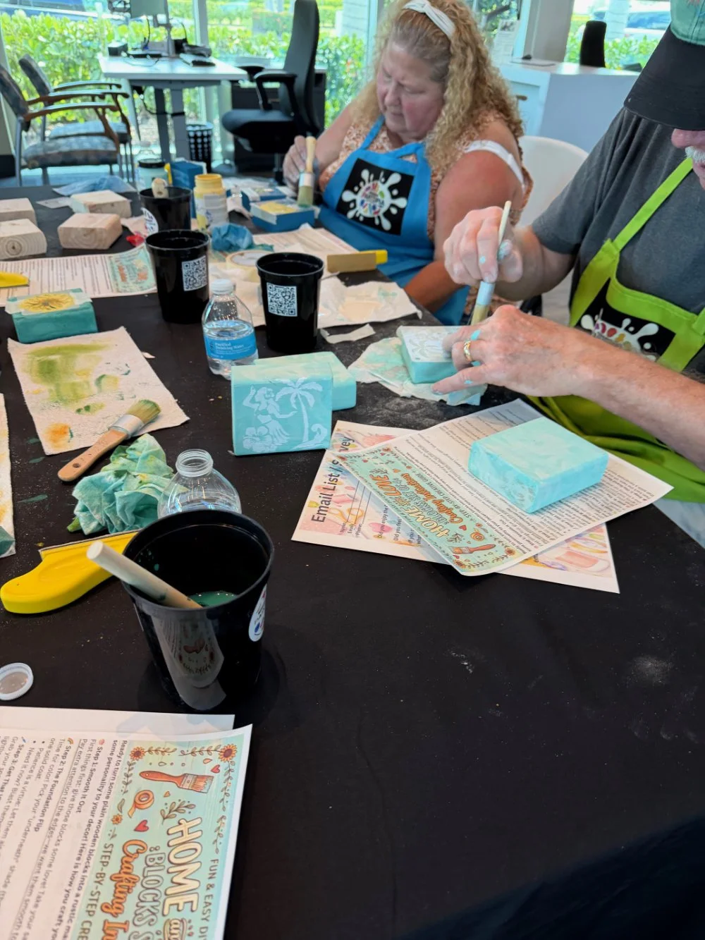 Two people working on creating soap art at a table with various tools, soap blocks, and printed instructions, in a well-lit room with a window view outside.