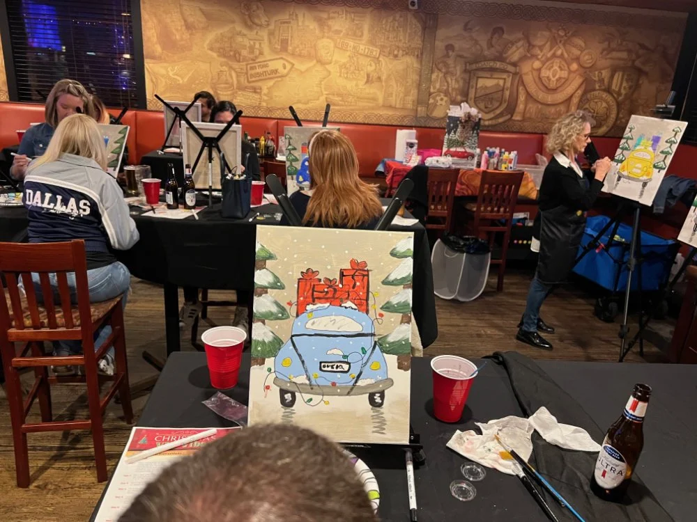 People painting at a holiday-themed painting class in a dimly lit room with a large wall mural. One painting features a blue car with Christmas presents on top surrounded by snow and trees.