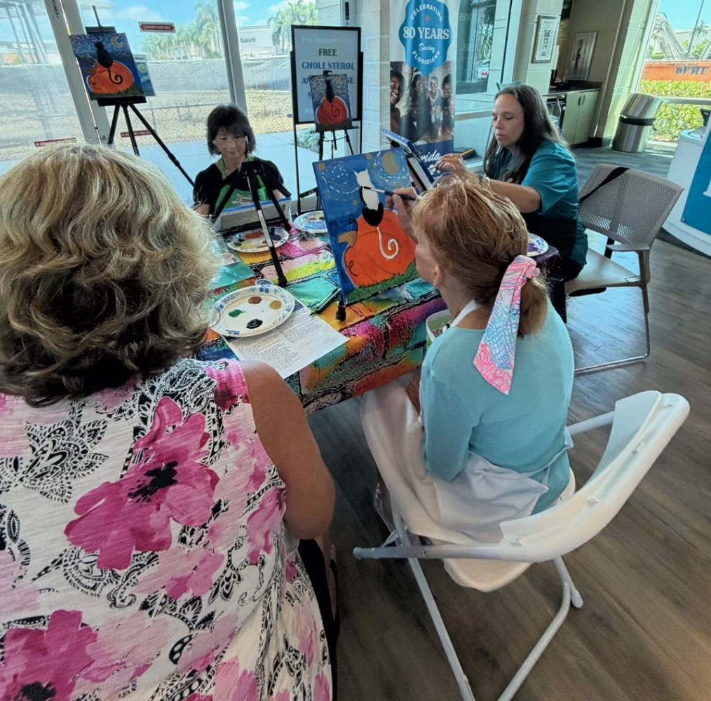 A group of women participating in a painting activity at a table in an indoor space with large windows. One woman is demonstrating painting a pumpkin with a black cat on it, while others watch and paint. Painting supplies and a palette are on the tab