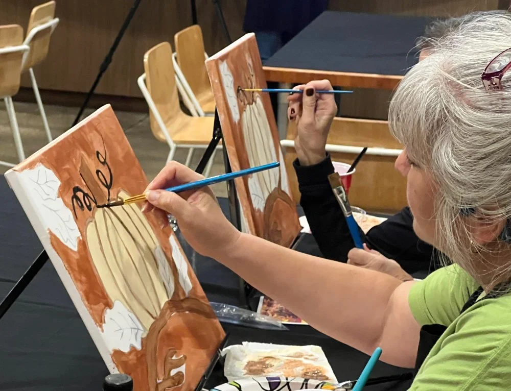 Two women painting fall-themed canvases featuring pumpkins and leaves at a group art event.