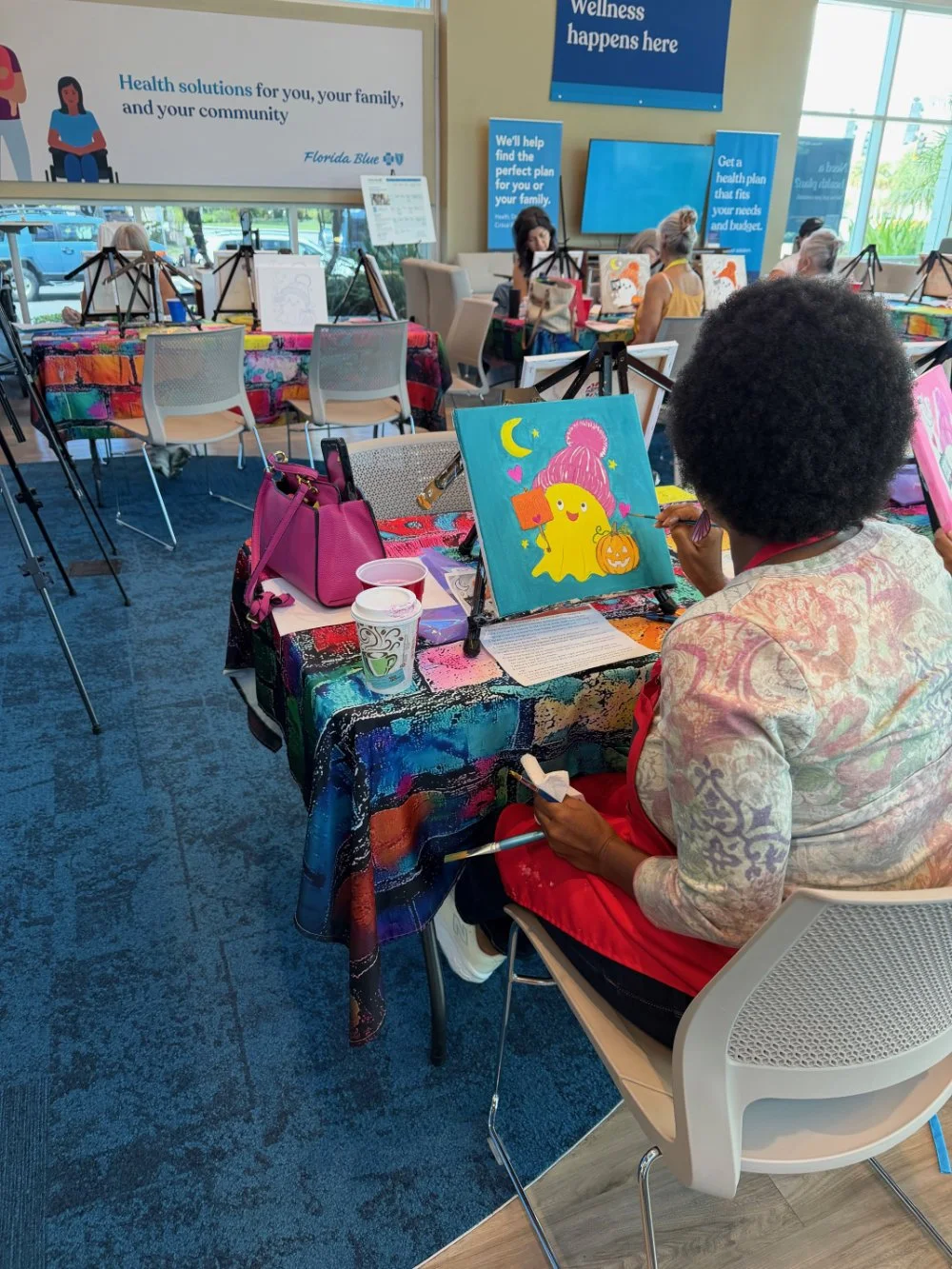 A woman with a short Afro hairstyle painting a colorful picture of a yellow blob character with a pink hat, holding a pumpkin-shaped bag in a well-lit indoor setting with others painting in the background.