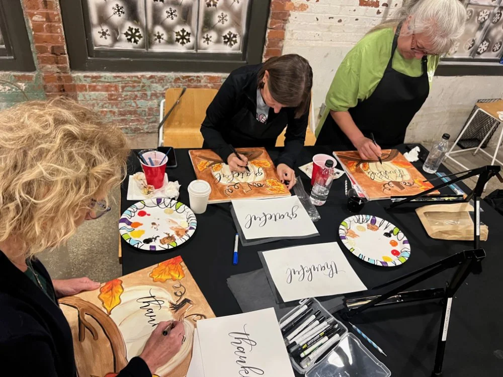 Three women painting fall-themed signs with the words 'Thankful', 'Blessed', and 'Grateful' on a table with art supplies, plates, and water bottles in a rustic indoor setting.