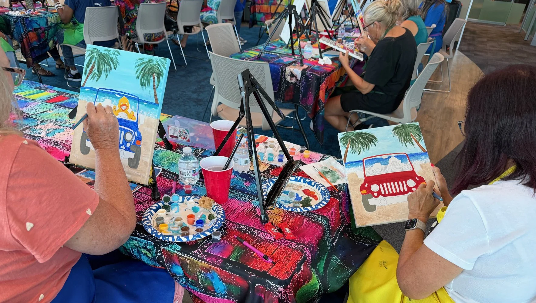 People painting beach scenes with red jeeps and palm trees at an indoor art class with colorful tablecloths and painting supplies.