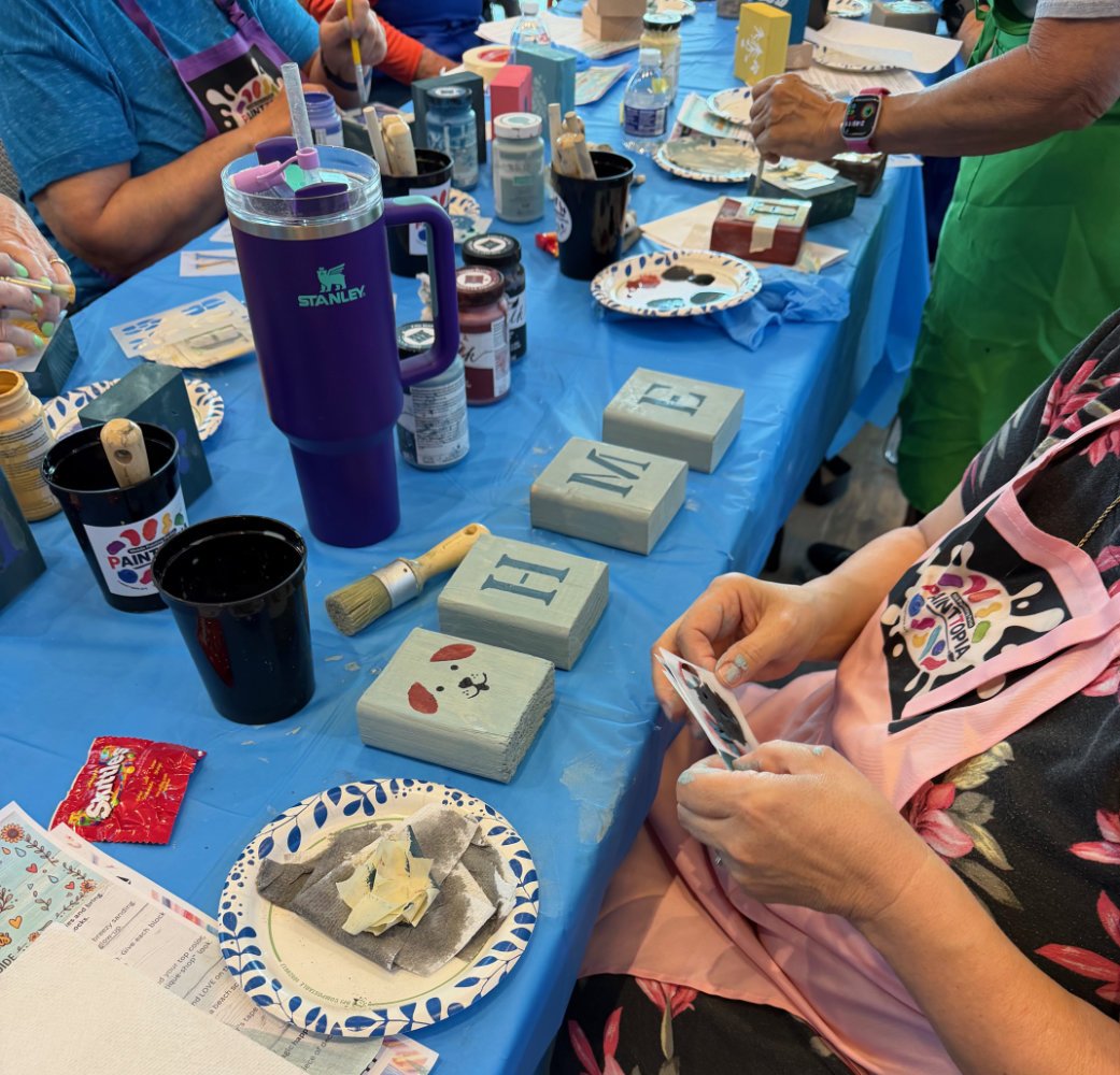 People participating in a craft activity, painting decorative blocks with letters and a dog face, on a blue tablecloth with supplies like paint, brushes, and paper plates.