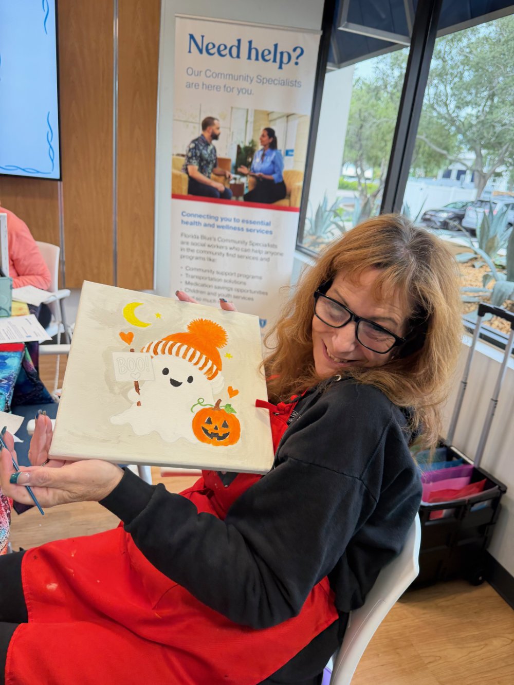 A woman with glasses and curly hair smiling and showing a Halloween-themed painted canvas featuring a friendly ghost with a striped orange hat, a pumpkin, and small hearts, in a community center or similar indoor space.