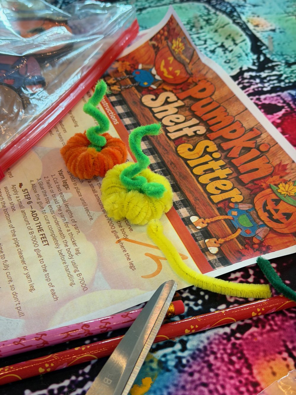 A colorful Halloween-themed craft project with small pipe cleaner pumpkins on a craft instruction sheet. An open plastic bag and scissors are also visible.