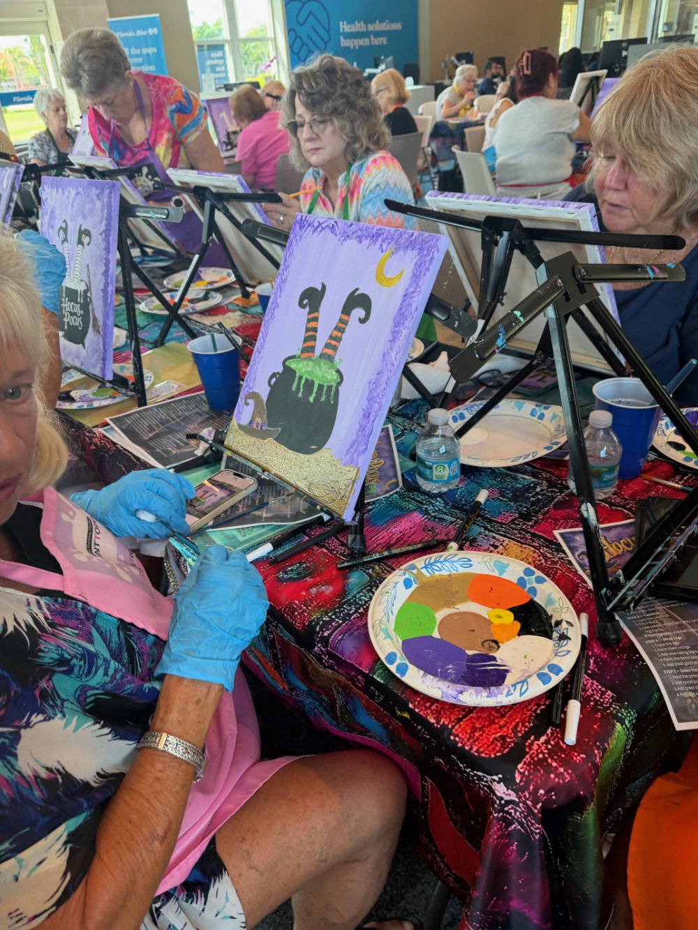 Group of women participating in a paint and sip event, painting Halloween-themed images, with canvases on easels, paint palettes, and drinks on a colorful table.