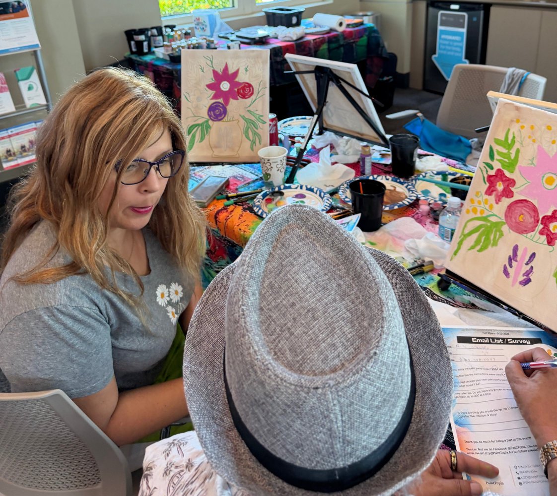 A woman with red hair and glasses sitting at a table with painting supplies, talking to a person wearing a wide-brimmed hat. The table has paintings, cups, and art materials, with colorful paintings and art supplies in the background.