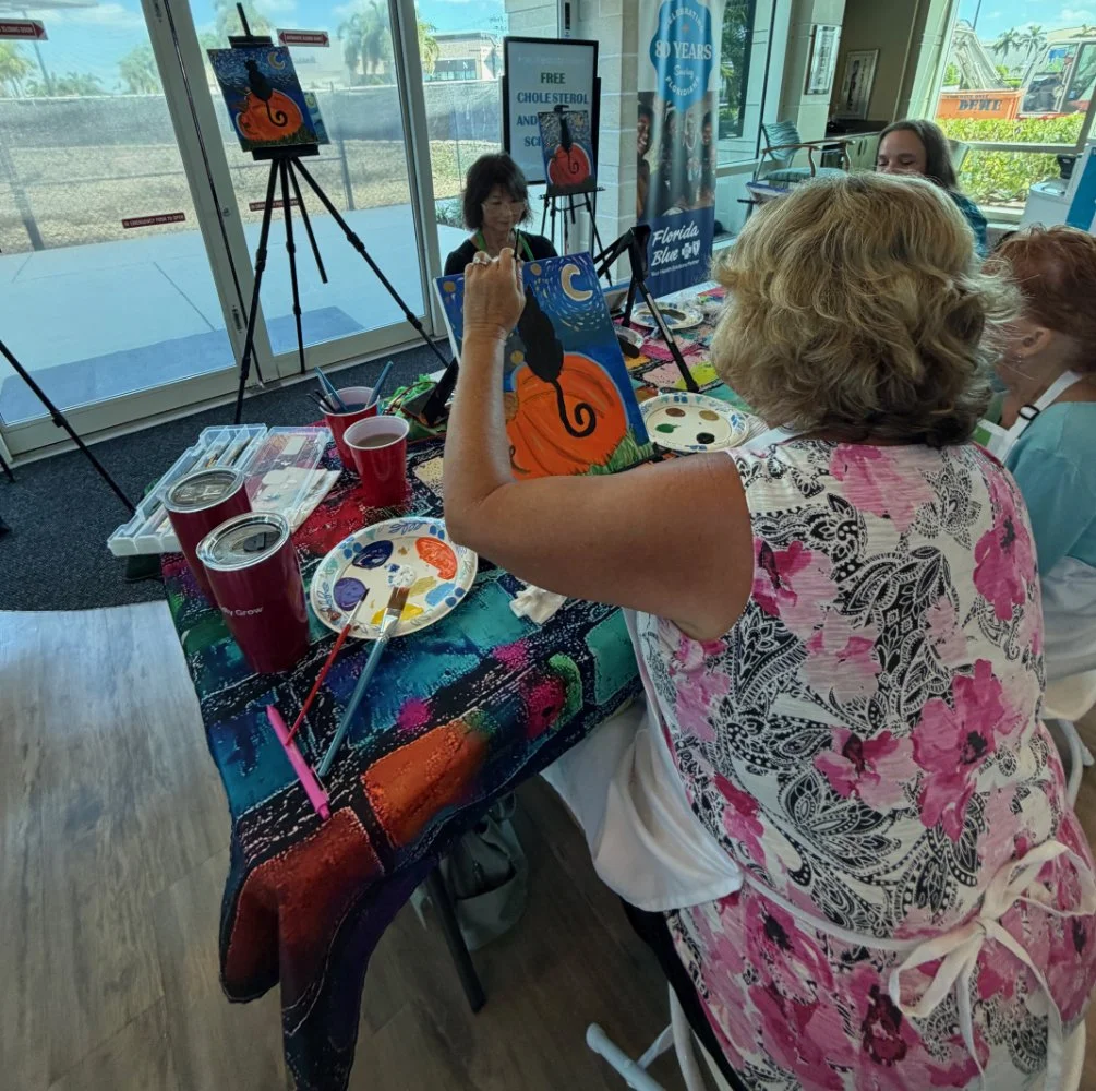 A woman painting a black cat on a pumpkin under a moonlit sky in a classroom or art studio. She is seated at a table covered with a colorful cloth, surrounded by paints, brushes, and cups of red-colored drinks. Several completed paintings of the same
