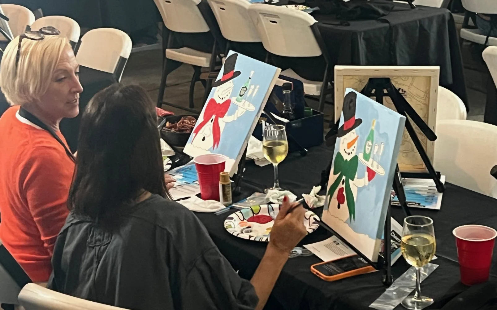 Two women sitting at a table painting snowman-themed artwork with easels, cups of wine, and art supplies on the table, in a social setting.