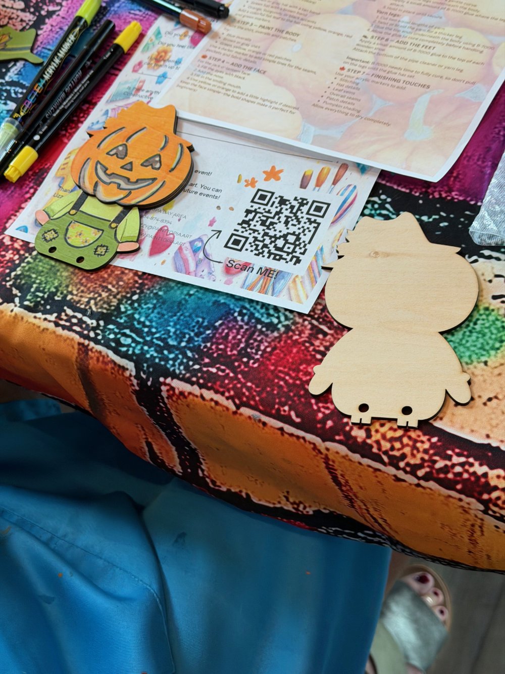 A Halloween-themed craft setup on a colorful tablecloth, including a pumpkin-shaped decoration, a blank wooden cutout of a character, colored pens, and a QR code on a paper.