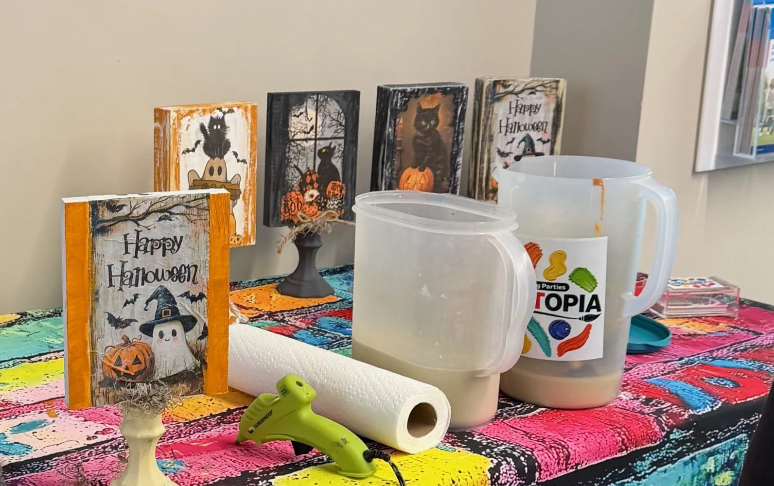 Table with Halloween decorations, tissue paper, and containers of slime. There are Halloween-themed signs and cutouts, including a ghost, black cat, pumpkin, and bats, with a colorful tablecloth.