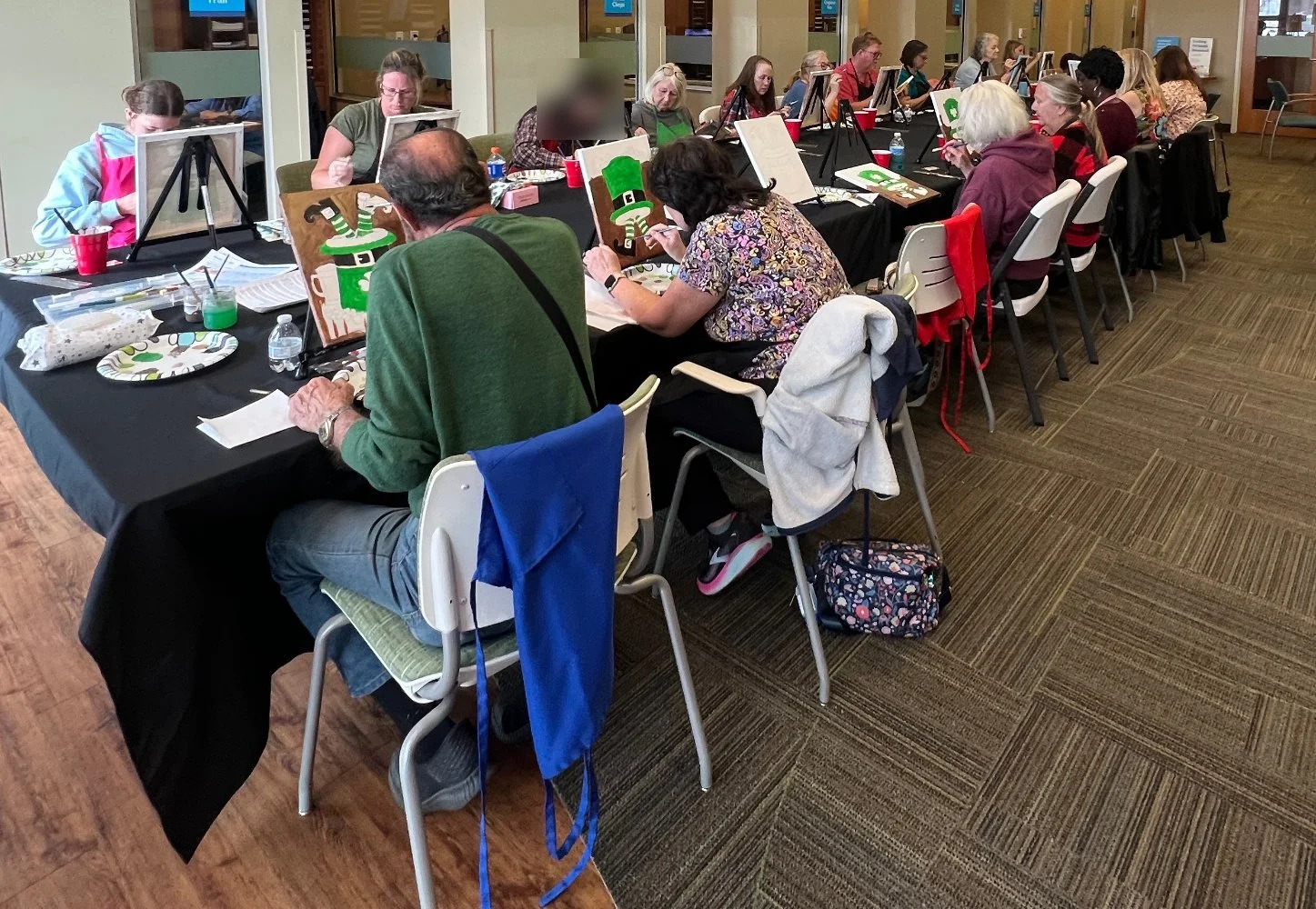Group of people painting holiday-themed pictures of green leprechauns at a long table in a community center or craft room.