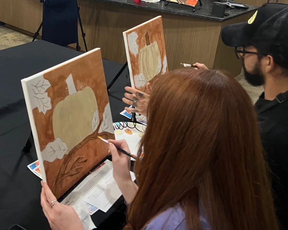 Two people painting autumn-themed canvases with pumpkins and leaves, sitting at a table in a room with black tablecloths.
