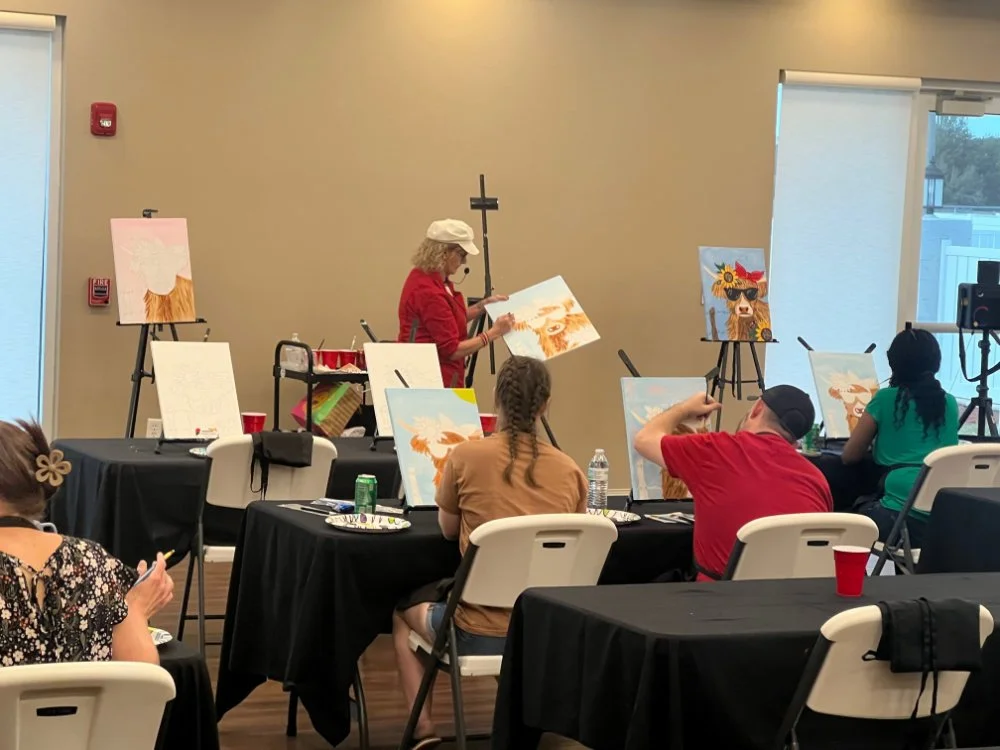 Art class with an instructor painting a lion on canvas, students seated at tables with paintings of lions, in a room with beige walls and large windows.