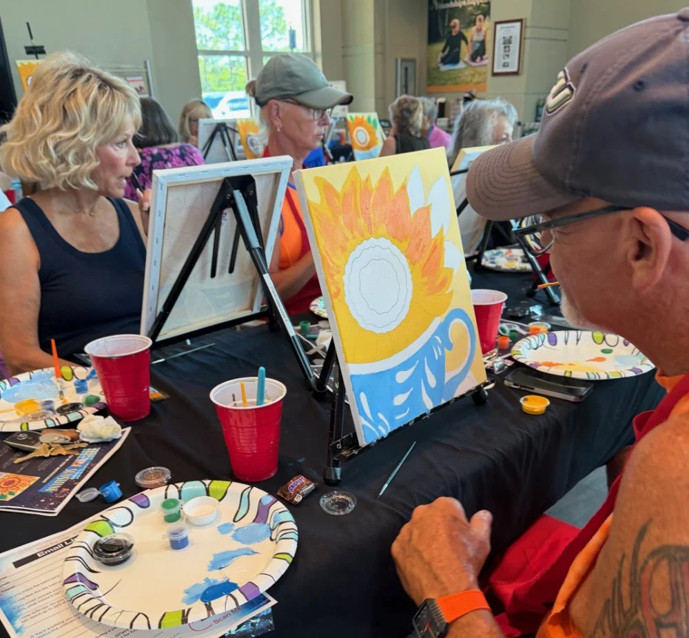 People participating in a paint-and-sip class, painting a sunflower on canvas, with art supplies and drinks on the table.